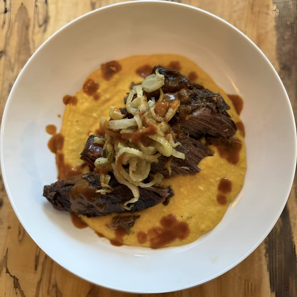Braised short ribs with caramelized onions and gravy over creamy polenta in a white bowl.